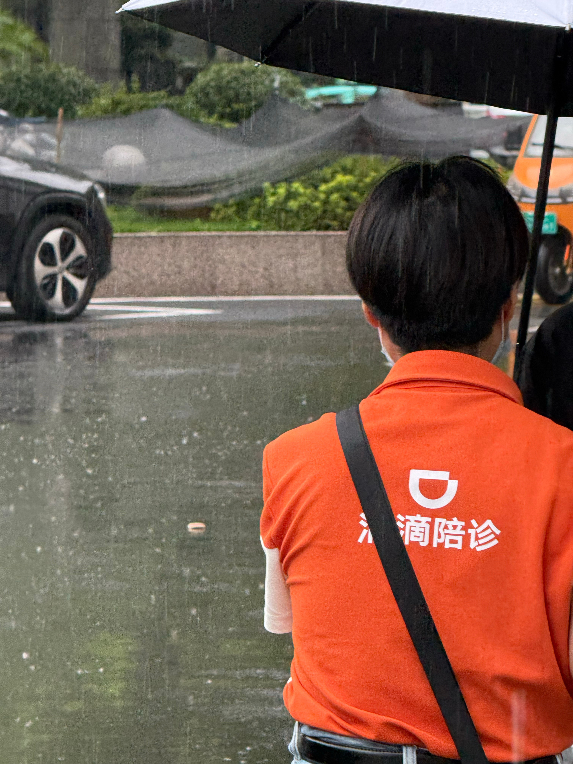 A Didi medical escort waits out the Hangzhou rain, the service's name — 滴滴陪诊 — printed across the back of the orange uniform. Photo: @张曼玉Mandy