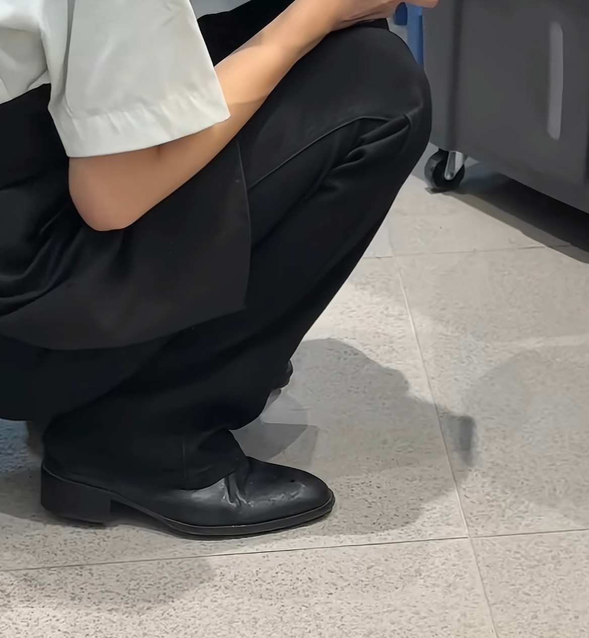 A worker in a white shirt and black trousers squats on a tiled floor, looking composed even in an informal moment.